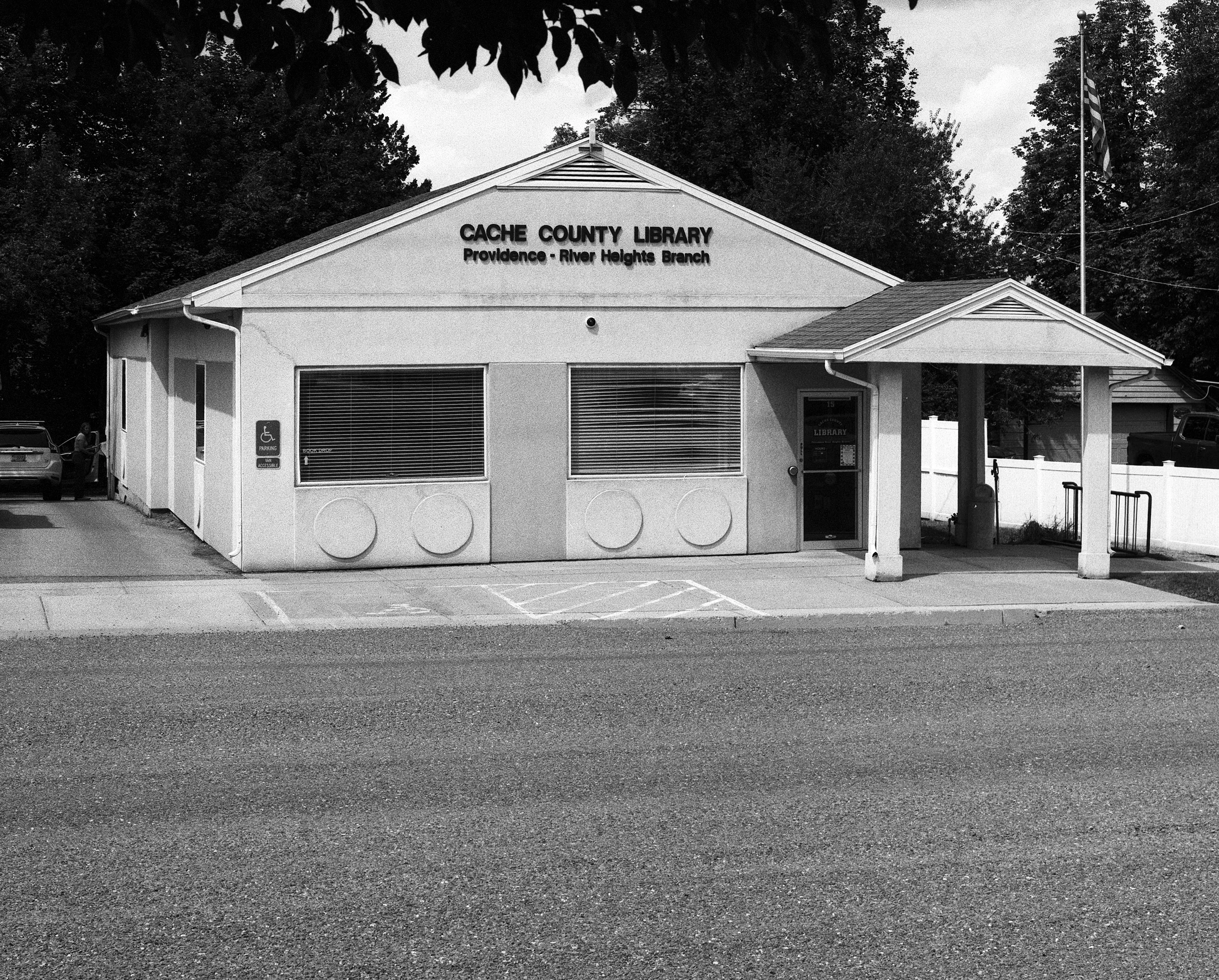 Black-and-white photo of the Cache County Library's Providence–River Heights Branch, a small single-story building with a covered entryway, framed by trees with an American flag flying nearby